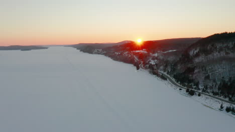 Vista-Aérea-De-Invierno-Sobre-Un-Río-Congelado-Mientras-El-Sol-Se-Pone-Detrás-De-Las-Colinas