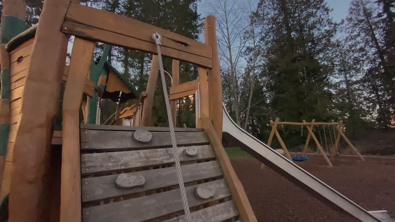 Childrens play equipment featuring rope pull climbing wall constructed from natural timber standing in neighborhood park under vibrant orange cloudy sunset sky captured via lateral camera sweep
