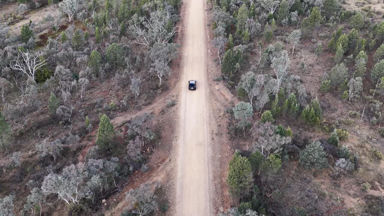 A single vehicle travels along a straight dirt road through sparse Australian bushland, captured from above with steady daylight and minimal camera movement
