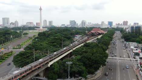 Schöne-Aussicht-Mit-Drohne-Auf-Das-Nationaldenkmal-Unter-Dem-Blauen-Himmel-Mit-Einem-Zug,-Der-In-Den-Bahnhof-Gambir-In-Jakarta,-Indonesien,-Fährt