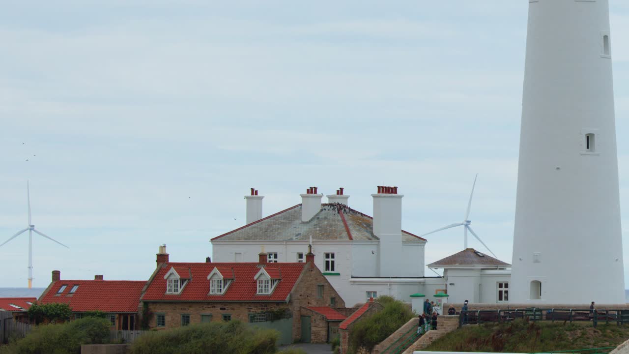 A stationary shot of a white lighthouse and red-roofed cottages on the Whitley Bay coastline, with wind turbines in the background under overcast daylight