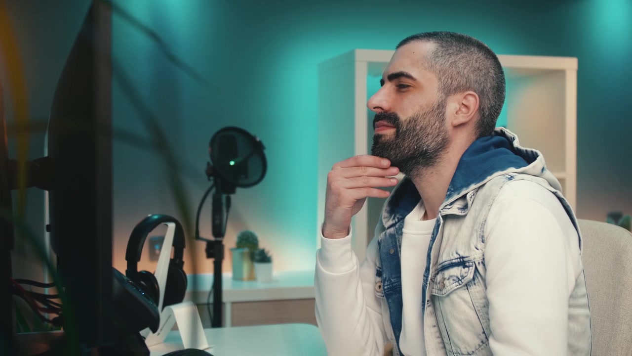 Giovane uomo con barba e capelli corti lavora diligentemente al computer in un colorato studio domestico. Ha un monitor nero e strisce di luci LED che creano un'atmosfera moderna e originale