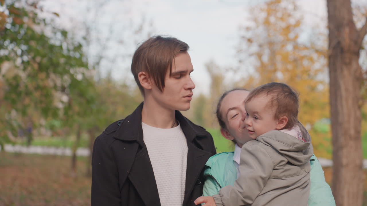 Giovani genitori caucasici in un parco autunnale, la madre culla il bambino baciandogli la guancia mentre il padre osserva con espressione pensierosa, foglie dorate, luce soffusa e nuvolosa, ritratto spontaneo, cappotti casual, emozionante.