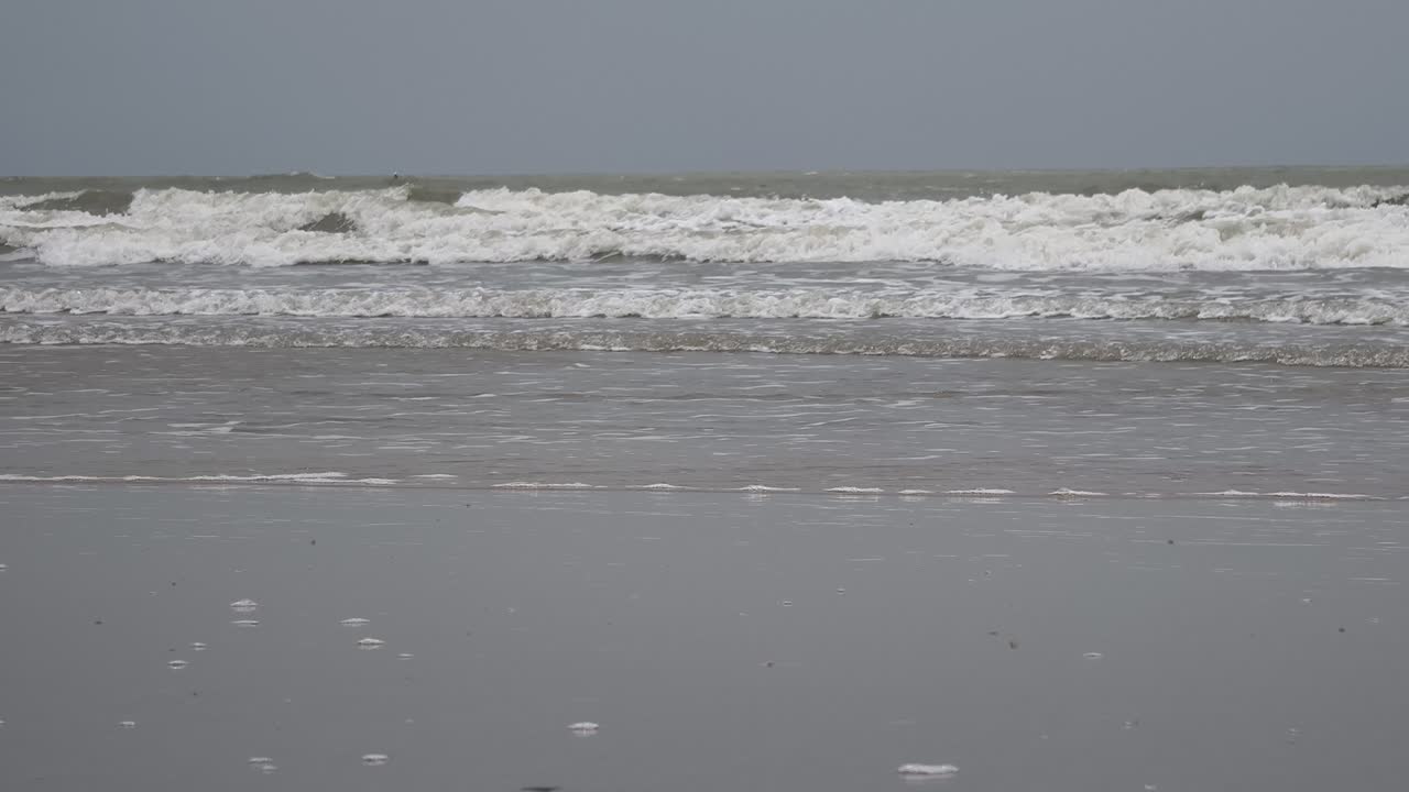 Un'ampia inquadratura di dolci onde oceaniche che si infrangono su una spiaggia sabbiosa bagnata durante una giornata nuvolosa in India. Adatto per vlog di viaggio, sfondi per la meditazione e narrazione ambientale.