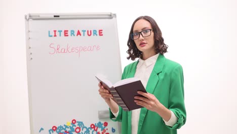 female teacher reading book by whiteboard with 'literature shakespeare'