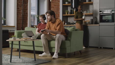 girl using laptop and boy playing video games sitting on sofa while their female roommate cleaning the house