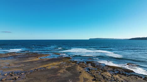 Aerial-flyover-of-rocky-ocean-coastline-on-east-coast-of-Australia,-low-angle-of-crashing-waves-on-beautiful-Longreef-shoreline,-Sydney