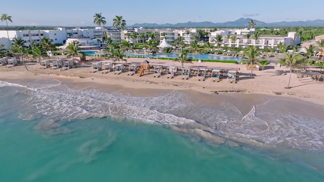 vista aérea que muestra el agua turquesa del mar del caribe, la lujosa playa de arena y el noble complejo nickelodeon, punta cana - modernas habitaciones de apartamentos blancos y cordilleras en el fondo