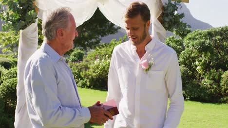 smiling caucasian senior male wedding officiant holding book and groom standing in outdoor altar