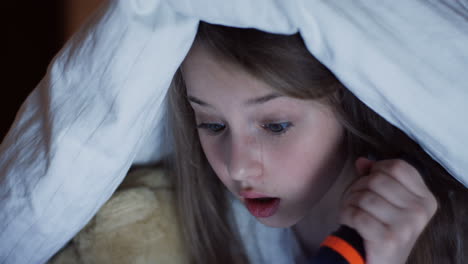 close-up view of cute little girl using a flashlight staring with surprised expression something under the blanket at night