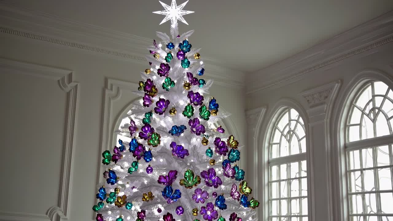 White Christmas tree decorated with colorful ornaments and a bright star, placed in a classic white room with large arched windows, creates a festive and elegant atmosphere