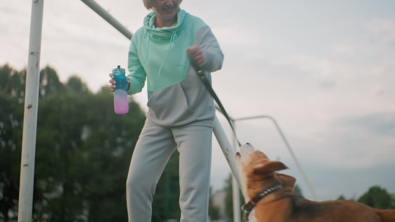 Elegant woman in joggers carrying water bottle swings leather leash playfully as energetic puppy jumps for it outdoors on grassy field during bright day capturing joyful bond fun