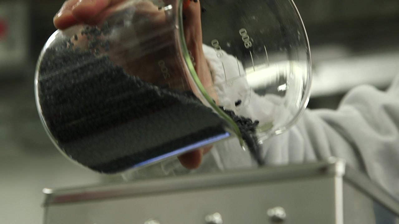 Technician pouring black plastic pellets into laboratory processing equipment before polymer extrusion and material testing in an industrial research environment.