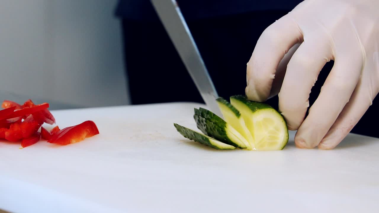 Chef's hands cutting a fresh cucumber. Hands in gloves cut cucumbers on the kitchen table. Cutting vegetables with a sharp knife. Close-up