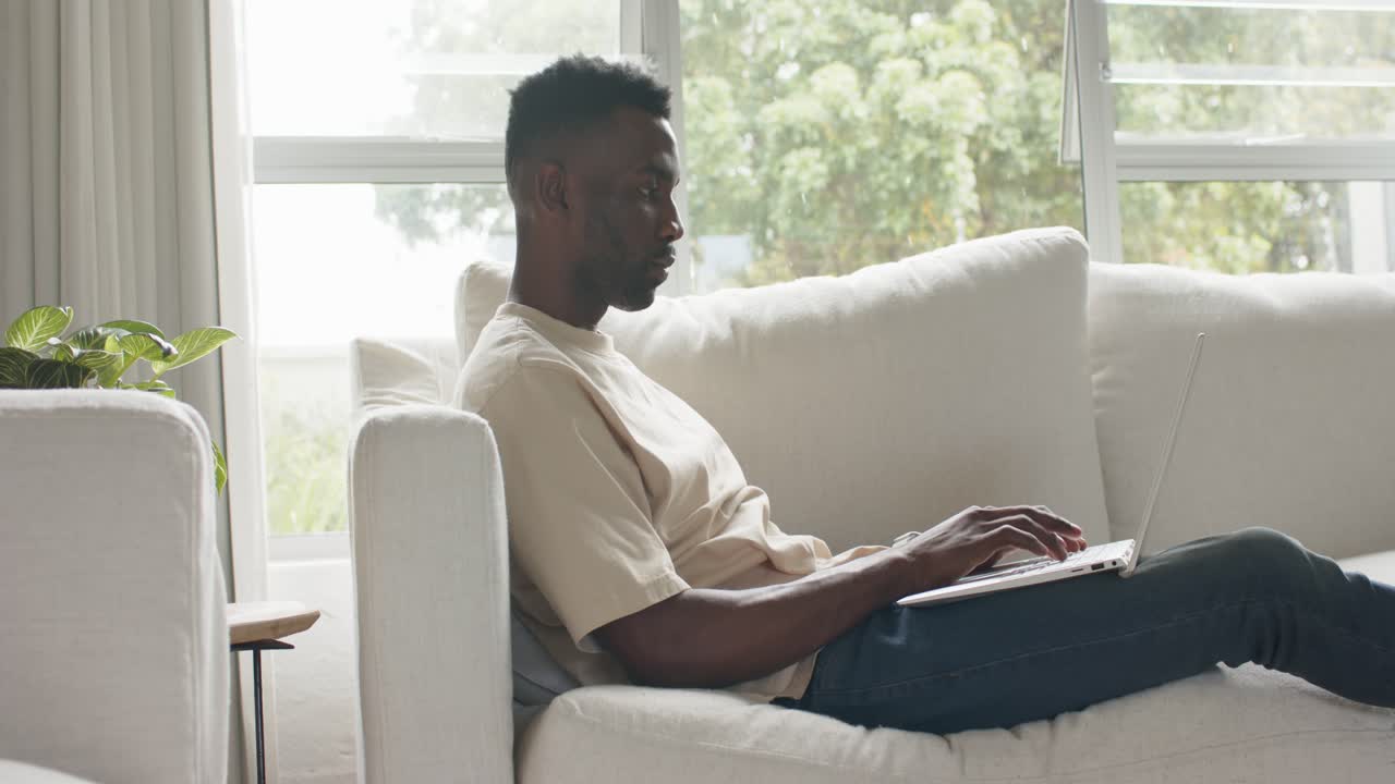 African adult male wearing beige T-shirt pausing typing to reach laptop trackpad on sofa for work
