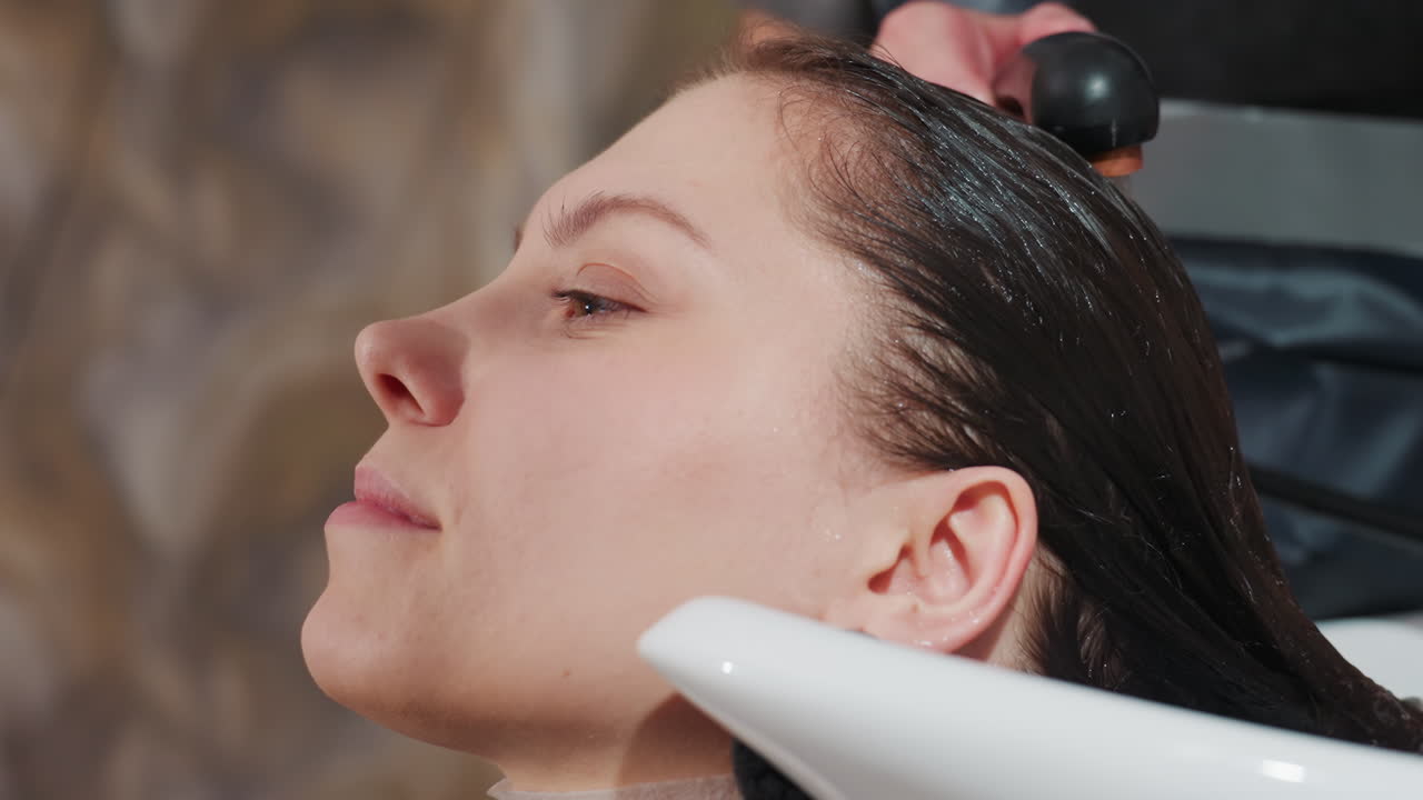 백인 여성이 미용실 세면대에서 부드러운 물로 헹굼을 즐기고 있어요. 노즐에서 나오는 물줄기가 어두운 머리카락의 샴푸를 헹궈내고 있으며, 목 쿠션이 보이고, 일정한 물줄기가 흐르고, 전문 스타일리스트가 온도와 압력을 조절하고 있어요.