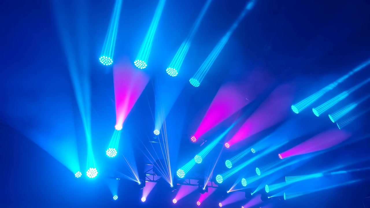 Bright moving beams sweep across a dark indoor venue with haze, creating a dramatic nightclub atmosphere with vivid blue, purple, and magenta lighting