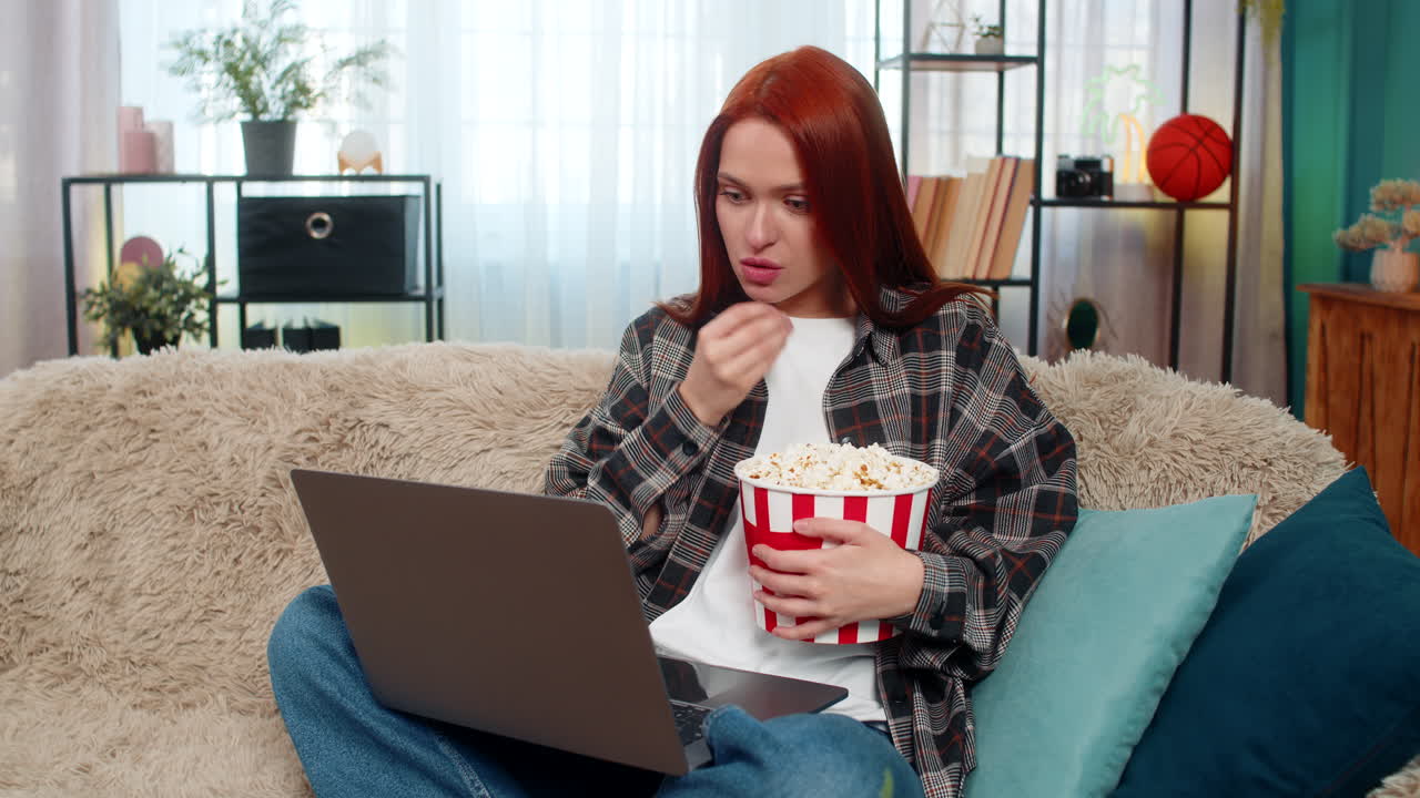 Woman sitting on sofa watching movie on laptop eating popcorn laughing emotionally with excitement
