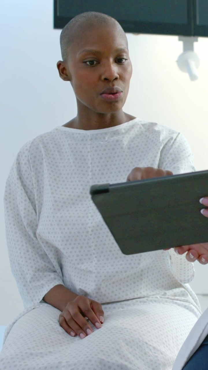 Vertical video: Reading patient reviewing tablet for care held by doctor on exam table wearing gown