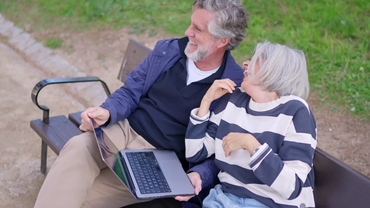 Pareja de ancianos felices riendo con un portátil en un banco del parque