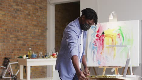 African-american-male-artist-wearing-face-mask-using-laptop-at-art-studio