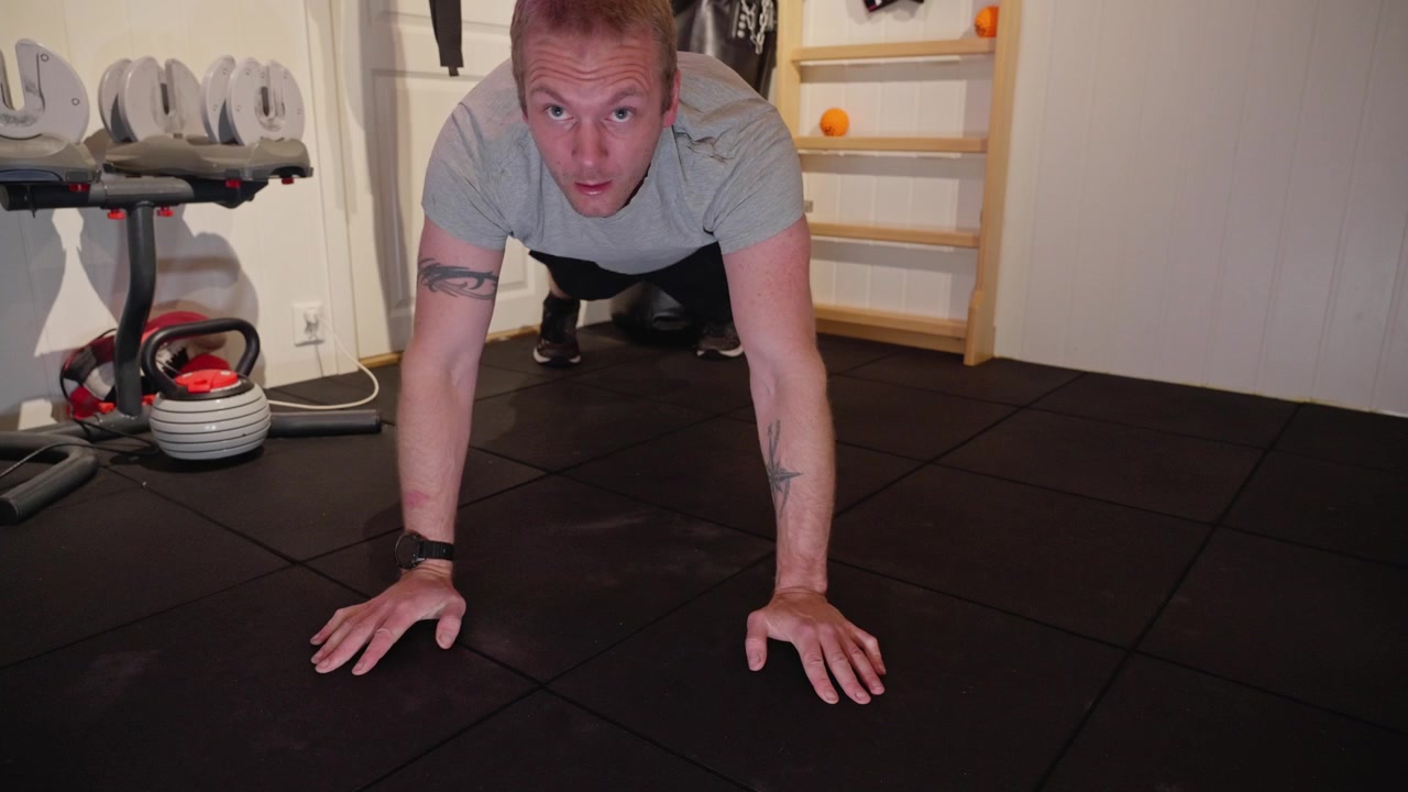A man in a home gym performs a plank to push up exercise, alternating arms while building strength and endurance