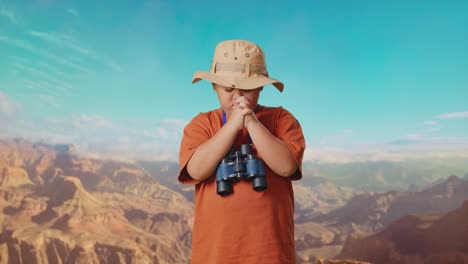 asian boy with a hat and binoculars praying for something while traveling at the top of mountain. boy researcher examines something, travel tourism adventure concept