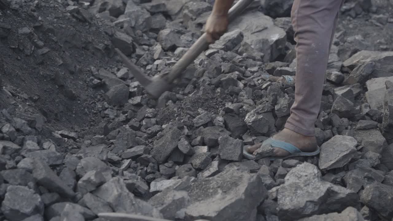 Closeup of a daily wage worker extracting coal at coal mine, wearing open toed sandal and pant, the individual is actively engaged in breaking or moving jagged pieces of coal using a hand tool