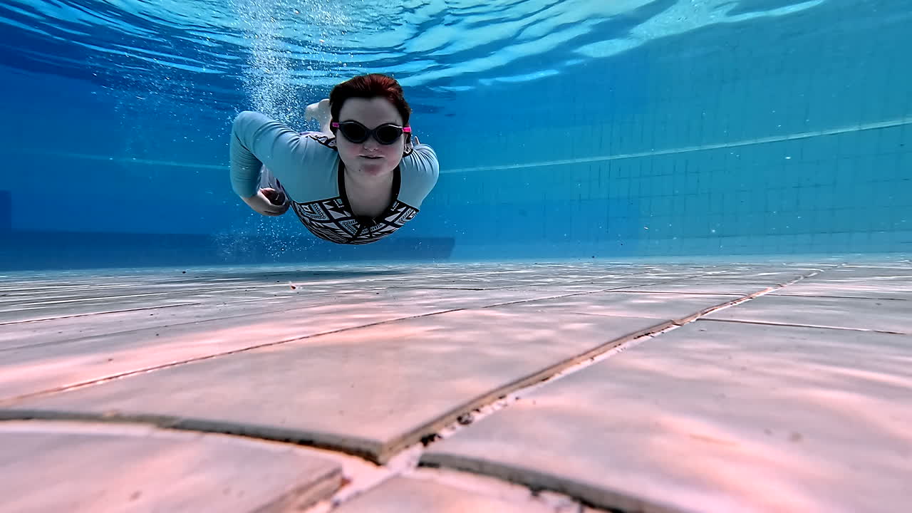 Young girl dives underwater, waves happily giving a thumbs up in crystal pool