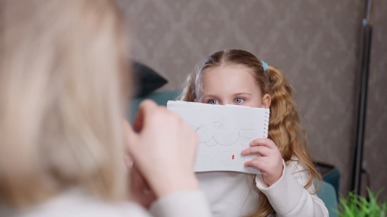 Young girl with long blonde hair in ponytails wearing white turtleneck showing drawing on paper to woman sitting across table indoors, focused expression, creative activity, family interaction in cozy home