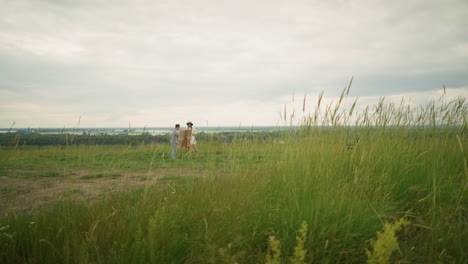 in a green field of tall grass, a man in a black hat and plaid shirt stands close to a woman in a white dress. they appear engaged in an observation, with a drawing board set up before them