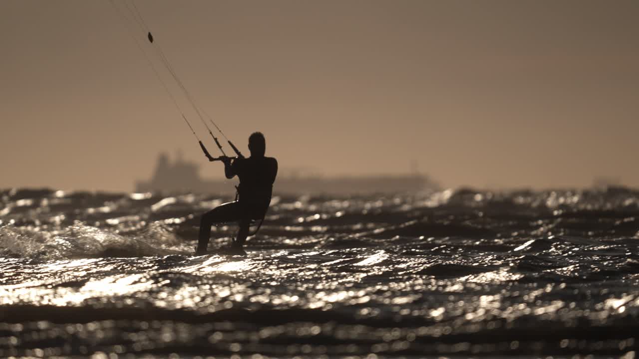 Bidikan menengah seseorang bersiluet yang sedang kitesurfing di lautan berombak dengan kapal di latar belakang menjelang matahari terbenam