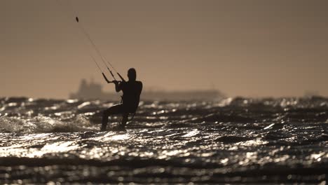 tiro medio de una persona con silueta de kitesurf en el océano agitado con un barco en el fondo cerca de la puesta del sol