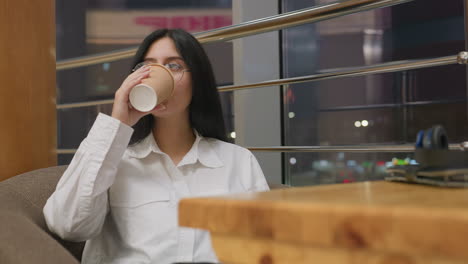 lady in glasses seated on cushioned chair near window calmly sips coffee with relaxed expression, enjoying quiet moment indoors with soft lighting and reflections from glass panel in modern setting