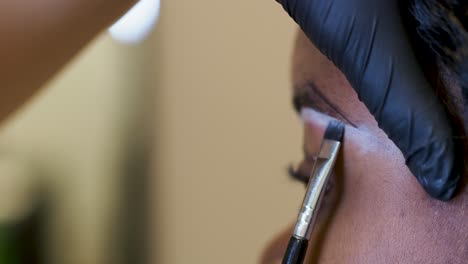 beautician is preparing a young black woman for a permanent tatoo session by drawing white correction lines above her eyebrows