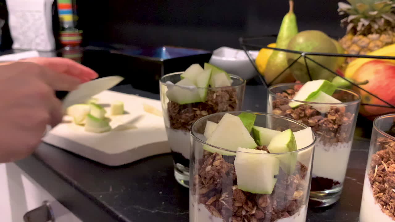 그 소녀는 손에 칼을 들고 나무 판자에 배를 자르고, 그녀 옆에는 요구르트와 플레이크 잔이 있습니다