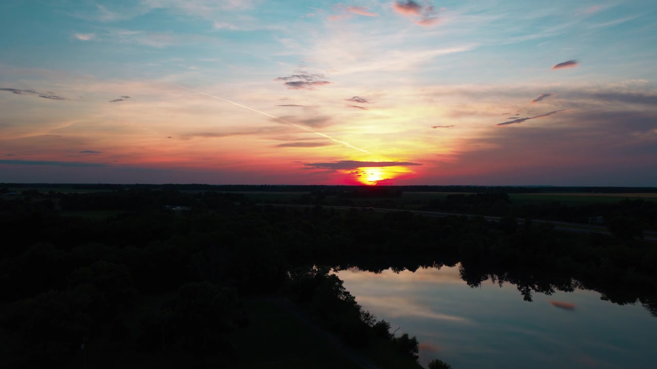 Vvid sunset over rural Richview, Illinois, the fiery sky and silhouetted treeline mirrored in a calm pond as the sun sets on the horizon.