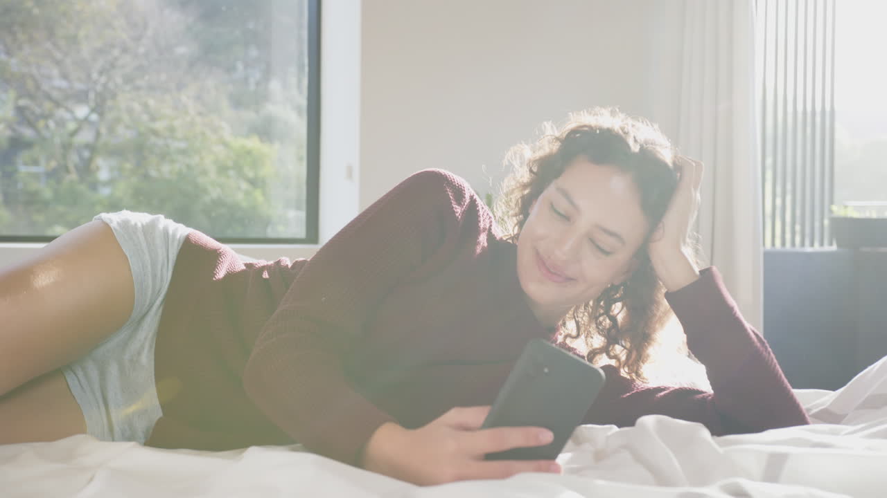 Relaxing on bed, woman smiling and using smartphone in bright room