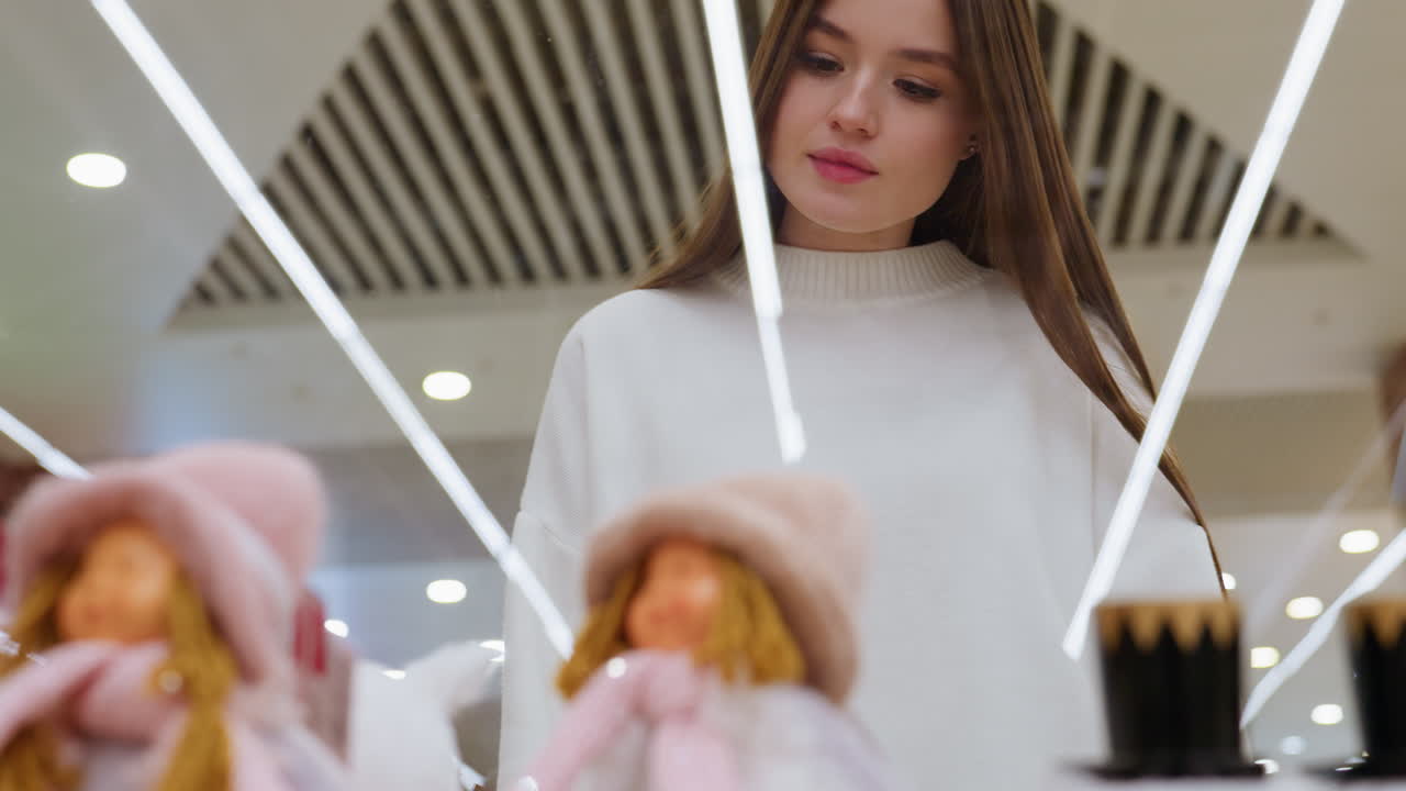 Young woman standing in front of decor store window, admiring dolls closely in a well-lit mall, reflections and vibrant mall lights visible with soft focus on background shoppers