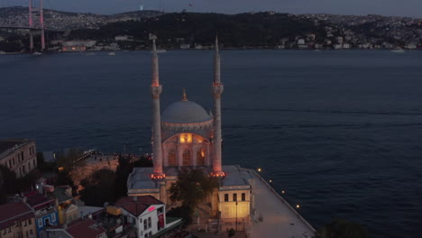 Ortakoy-Mosque-illuminated-in-beautiful-yellow-light-on-Water-side-at-Dusk-with-Bosphorus-and-Bridge,-Aerial-wide-shot