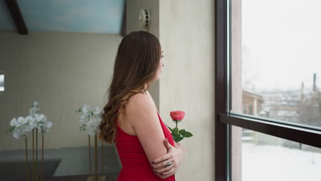 mujer observa el invierno fuera de la ventana. mujer modelo en elegante vestido rojo con rosa en la mano refleja en el mundo nevado exterior con sentido de introspección