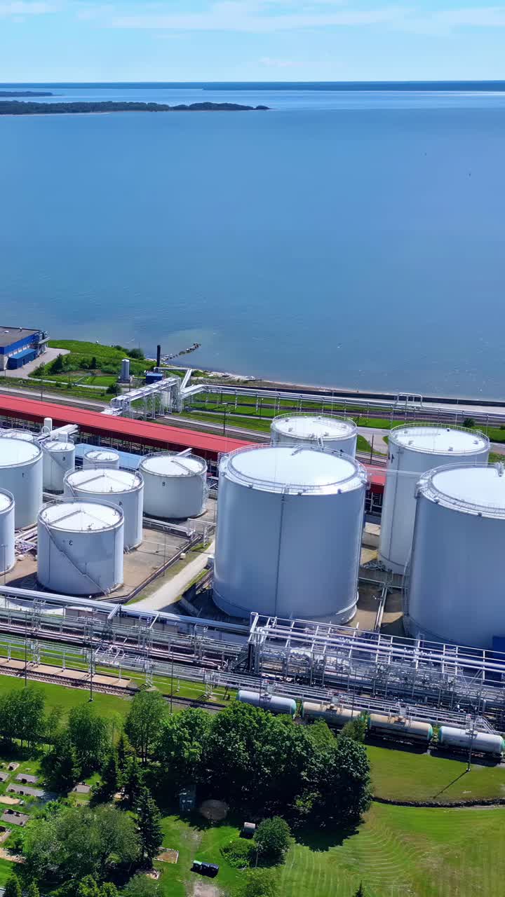 Aerial shot showcasing a modern oil or chemical terminal in Ventspils, Latvia, featuring numerous white storage tanks, pipelines, and a serene waterfront