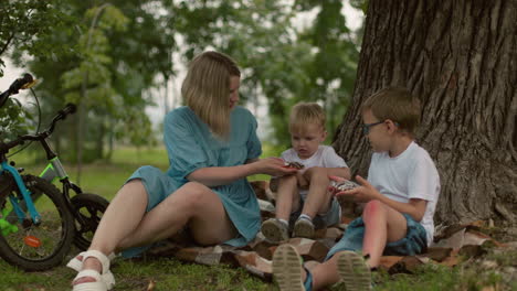 a mother sits on the ground with her two children under a tree, offering something to the younger child, who refuses it