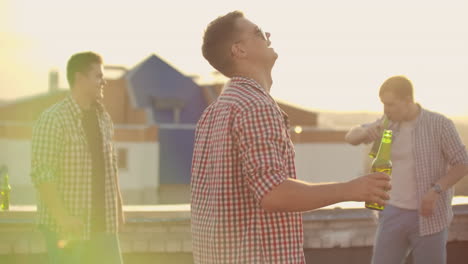 A-young-American-man-in-trendy-glasses-with-beer-moves-in-a-dance-at-a-party-with-his-friends-on-the-roof.-He-smiles-and-has-fun.