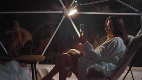 woman examines glass of alcoholic drink under lighting of lamp. female sits near panoramic windows overlooking nightlife in dimly lit hotel room for tourists