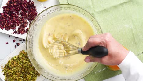 baking a dessert called persian love cake made with rose petals and pistachios. cake with white glaze frosting. using a whisk to combine ingredients. beautiful cake for tea time or celebration