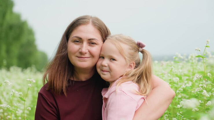 Weiße Mutter und Tochter umarmen sich auf einer Wildblumenwiese, eine Nahaufnahme, die die liebevolle Bindung einfängt, die Mutter tröstet und küsst das Kind, sanftes Lächeln, üppiges grünes Laub, zarte weiße Blüten, weich