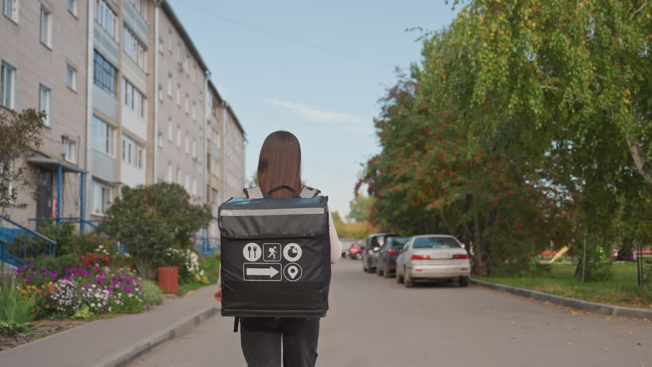 En kurér med rygsæk går ned af en forstadsgade forbi parkerede biler og lejlighedsbygninger med sommerligt løv og en rolig atmosfære. Kuréren nærmer sig en dør og tjekker sin telefon for at finde vej.
