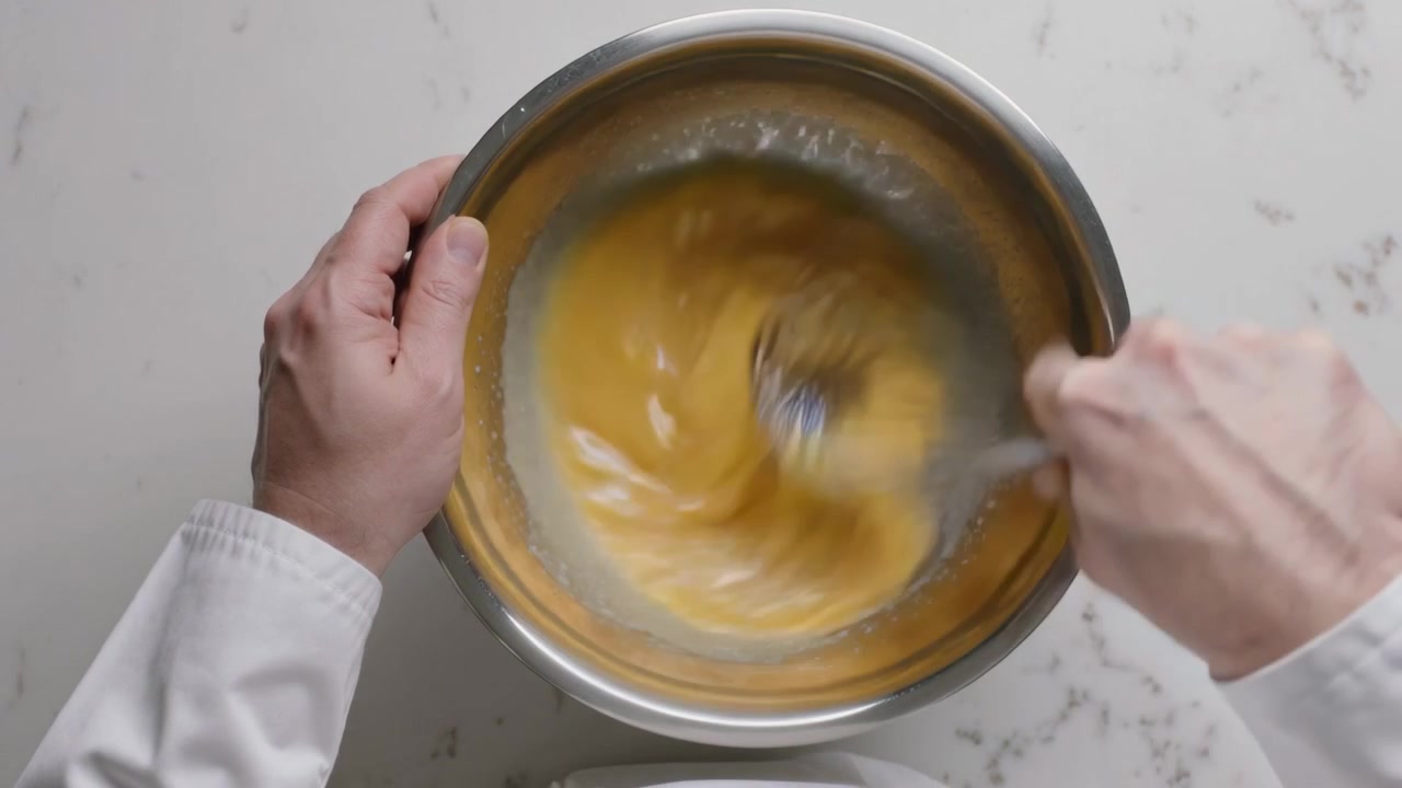Mescolare la pastella in una ciotola d'acciaio sul bancone, mano destra che usa la frusta per mescolare, maniche bianche che stabilizzano.