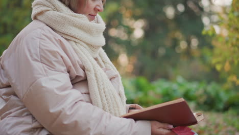 close up side view of mature woman wrapped in cozy scarf focusing on open book while gently holding red autumn leaf in hand, soft bokeh forest background